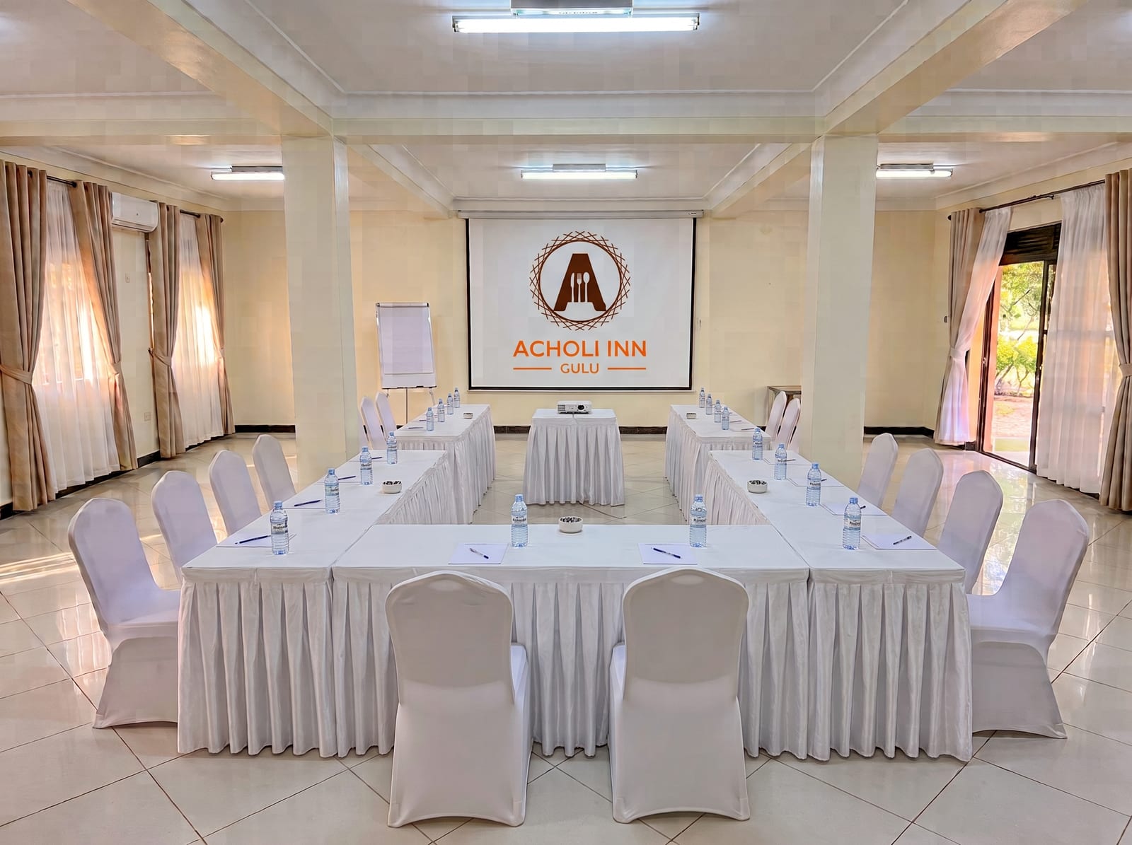 Acholi Inn meeting room set up with a U-shaped table, white chairs and the Acholi Inn logo on the projector screen
